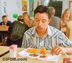 A Man Is Sitting At A Table Eating A Tray Of Food In A Cafeteria . GIF