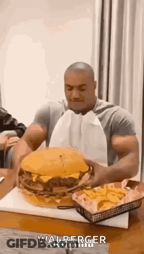 A Man Is Sitting At A Table Eating A Very Large Hamburger And French Fries . GIF