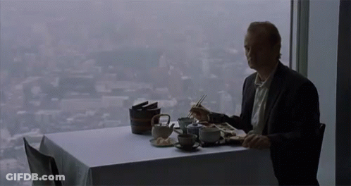 A Man Is Sitting At A Table Eating Food With Chopsticks And Looking Out A Window . GIF