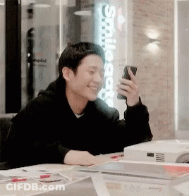 A Man Is Sitting At A Table Holding A Cell Phone And Smiling GIF