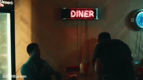A Man Is Sitting At A Table In A Diner With A Neon Sign Above Him . GIF