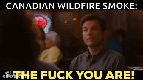 A Man Is Sitting At A Table In A Restaurant With A Sign That Says Canadian Wildfire Smoke The Fuck You Are . GIF