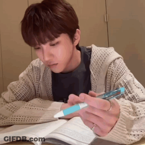 A Man Is Sitting At A Table Reading A Book And Holding A Pen GIF