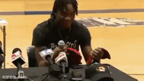 A Man Is Sitting At A Table Surrounded By Microphones Holding A Hat . GIF