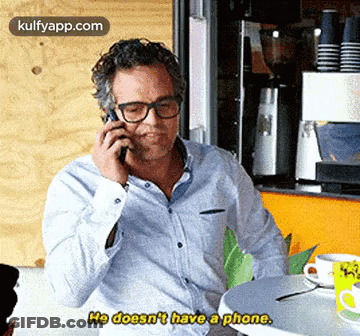 A Man Is Sitting At A Table Talking On A Cell Phone And Says He Doesn 'T Have A Phone . GIF