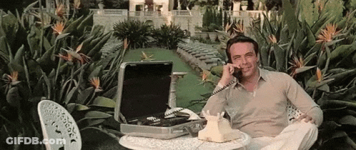 A Man Is Sitting At A Table Talking On A Telephone In A Garden . GIF