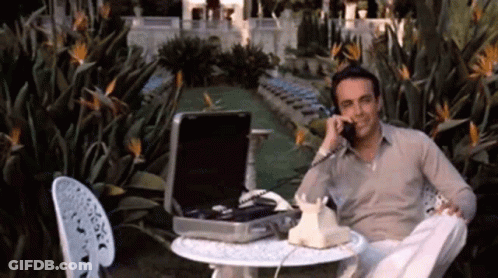 A Man Is Sitting At A Table With A Briefcase And Talking On A Phone GIF