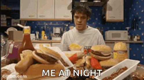 A Man Is Sitting At A Table With A Bunch Of Food And The Words Me At Night Are Above Him GIF