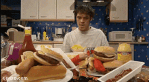 A Man Is Sitting At A Table With A Bunch Of Food Including Hamburgers And Hot Dogs .. GIF
