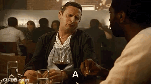 A Man Is Sitting At A Table With A Glass Of Wine And Talking To Another Man In A Restaurant . GIF