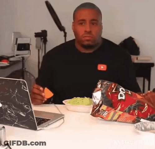 A Man Is Sitting At A Table With A Laptop And A Bag Of Doritos . GIF