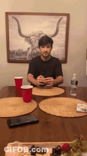 A Man Is Sitting At A Table With A Picture Of A Bull On The Wall Behind Him GIF