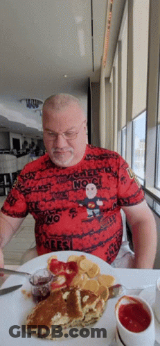 A Man Is Sitting At A Table With A Plate Of Food And A T-Shirt That Says " No " GIF