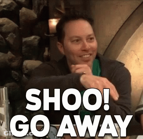 A Man Is Sitting At A Table With A Sign That Says Shoo Go Away On It . GIF