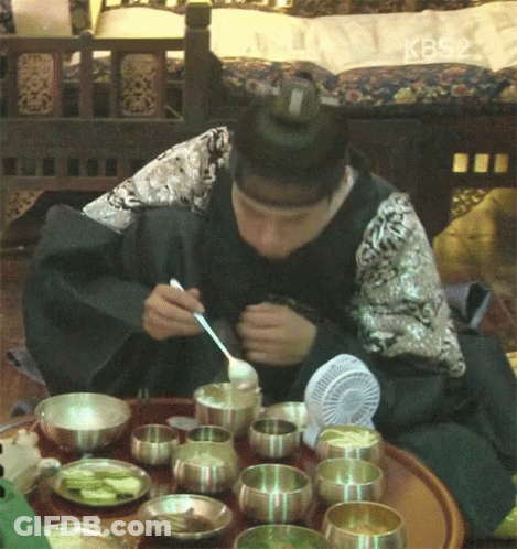 A Man Is Sitting At A Table With Bowls Of Food And A Fan And Eating With A Spoon And Fork GIF