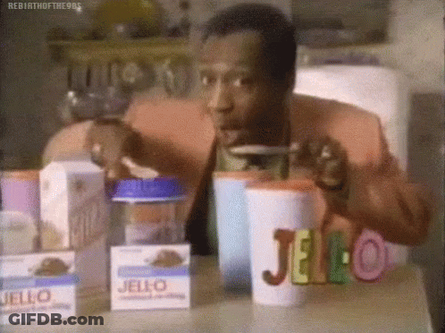 A Man Is Sitting At A Table With Boxes Of Jello And Drinking From A Cup . GIF