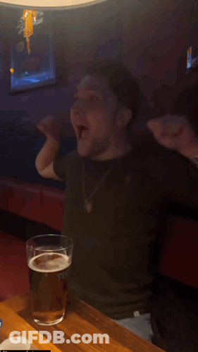 A Man Is Sitting At A Table With His Arms In The Air And A Glass Of Beer In Front Of Him That Says " The " GIF
