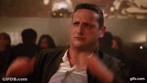 A Man Is Sitting At A Table With His Hands On His Chest In A Crowded Room . GIF