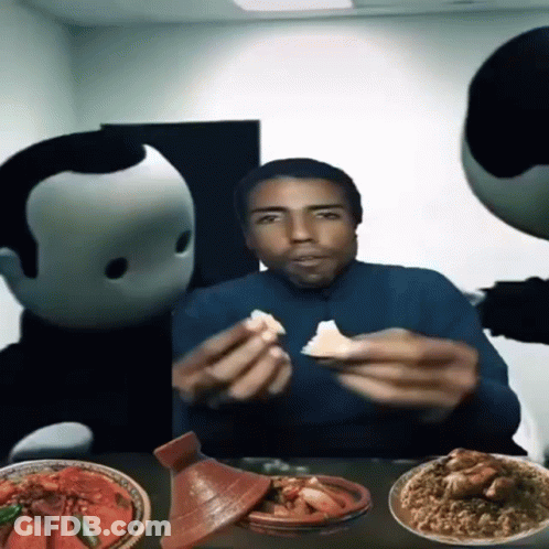 A Man Is Sitting At A Table With Plates Of Food And Two Mascots Behind Him GIF