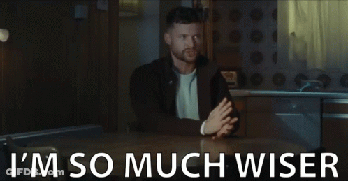 A Man Is Sitting At A Table With The Words `` I 'M So Much Wiser '' Written On The Bottom . GIF