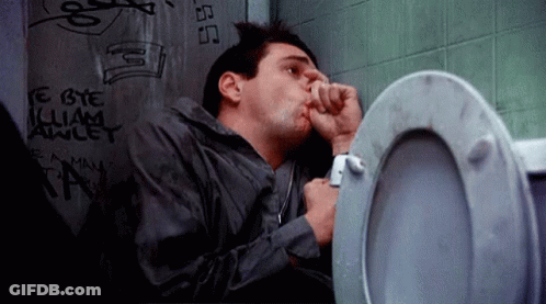A Man Is Sitting In A Bathroom Next To A Toilet With Graffiti On The Wall Behind Him . GIF