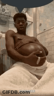 A Man Is Sitting In A Bathroom With A Towel Around His Waist And Holding His Belly . GIF