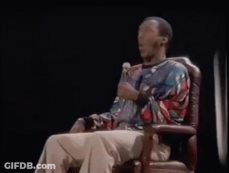 A Man Is Sitting In A Chair Holding A Microphone And Talking Into It . GIF