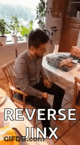 A Man Is Sitting In A Chair In Front Of A Table With A Stack Of Books And The Words Reverse Jinx On It . GIF