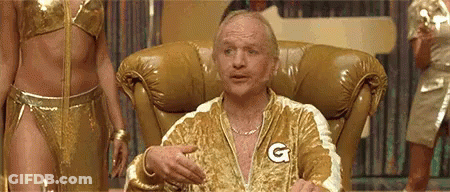 A Man Is Sitting In A Chair Wearing A Gold Jacket With A G On It . GIF