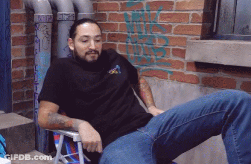 A Man Is Sitting In A Chair With A Brick Wall Behind Him With Graffiti On It That Says ' Rick ' GIF
