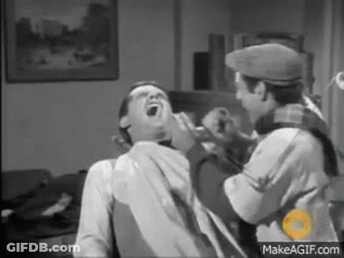 A Man Is Sitting In A Chair With His Mouth Open While Another Man Shaves His Beard . GIF