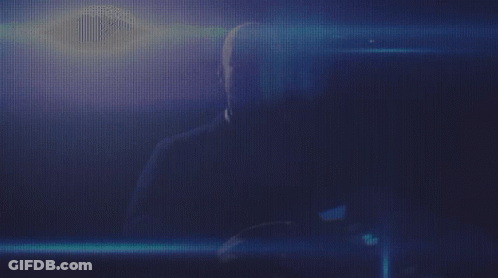 A Man Is Sitting In A Dark Room With A Blue Light Behind Him GIF