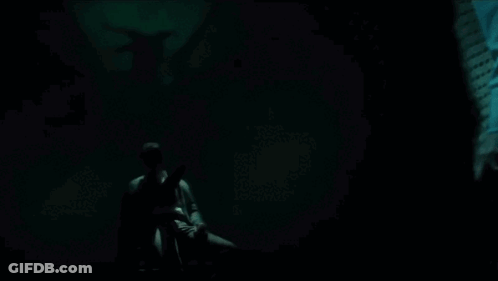 A Man Is Sitting In A Dark Room With A Statue Of A Demon In The Background . GIF