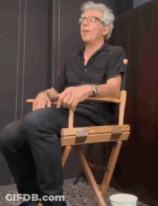 A Man Is Sitting In A Director 'S Chair With His Legs Crossed GIF