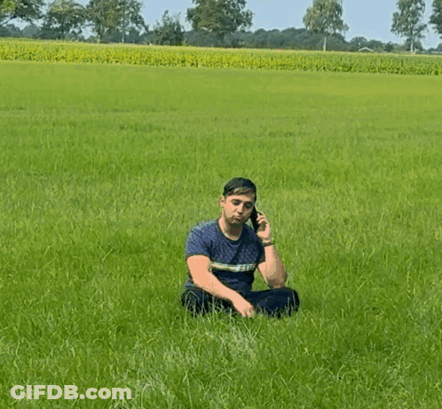 A Man Is Sitting In A Field Talking On A Cell Phone GIF
