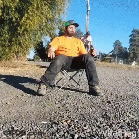 A Man Is Sitting In A Folding Chair On The Side Of A Road Holding A Bottle Of Beer . GIF
