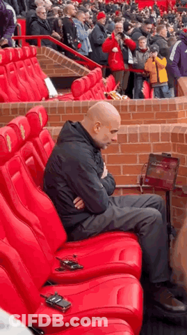A Man Is Sitting In A Row Of Red Seats In A Stadium With His Arms Crossed . GIF
