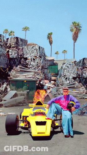 A Man Is Sitting In A Yellow Race Car With Flames Coming Out Of The Steering Wheel GIF