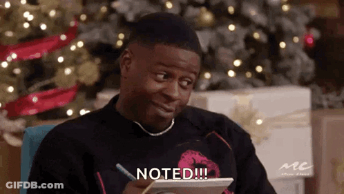 A Man Is Sitting In Front Of A Christmas Tree And Writing On A Piece Of Paper With The Words Noted Written On It . GIF