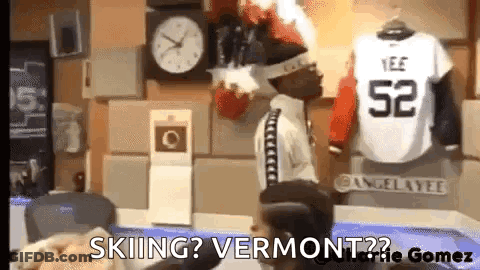 A Man Is Sitting In Front Of A Clock And A Jersey That Says ' Skiing ? Vermont ? ' GIF