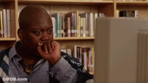 A Man Is Sitting In Front Of A Computer In A Library With His Hand On His Chin . GIF