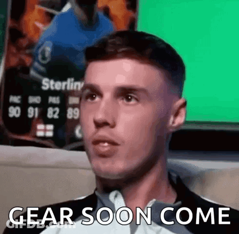 A Man Is Sitting In Front Of A Green Screen With The Words `` Gear Soon Come '' Written On It . GIF
