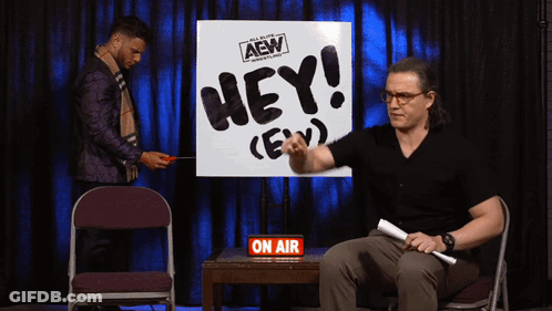 A Man Is Sitting In Front Of A Sign That Says Hey GIF