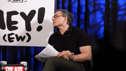 A Man Is Sitting In Front Of A Sign That Says Hey GIF