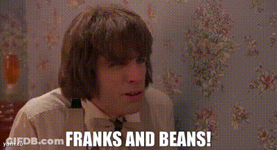 A Man Is Sitting In Front Of A Wall With Flowers On It And Says `` Franks And Beans '' . GIF