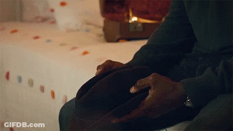 A Man Is Sitting On A Bed Holding A Brown Hat In His Hands . GIF