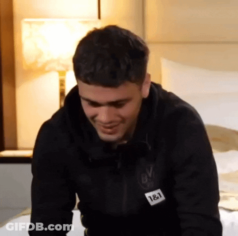 A Man Is Sitting On A Bed Wearing A Black Jacket With The Number 18 On It . GIF