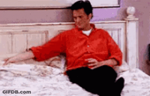 A Man Is Sitting On A Bed With A Drink In His Hand . GIF
