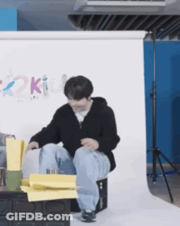 A Man Is Sitting On A Box With His Legs Crossed In Front Of A Wall With The Word Kit On It GIF