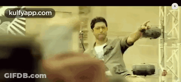 A Man Is Sitting On A Chair With His Arms Outstretched And Pointing At Something . GIF